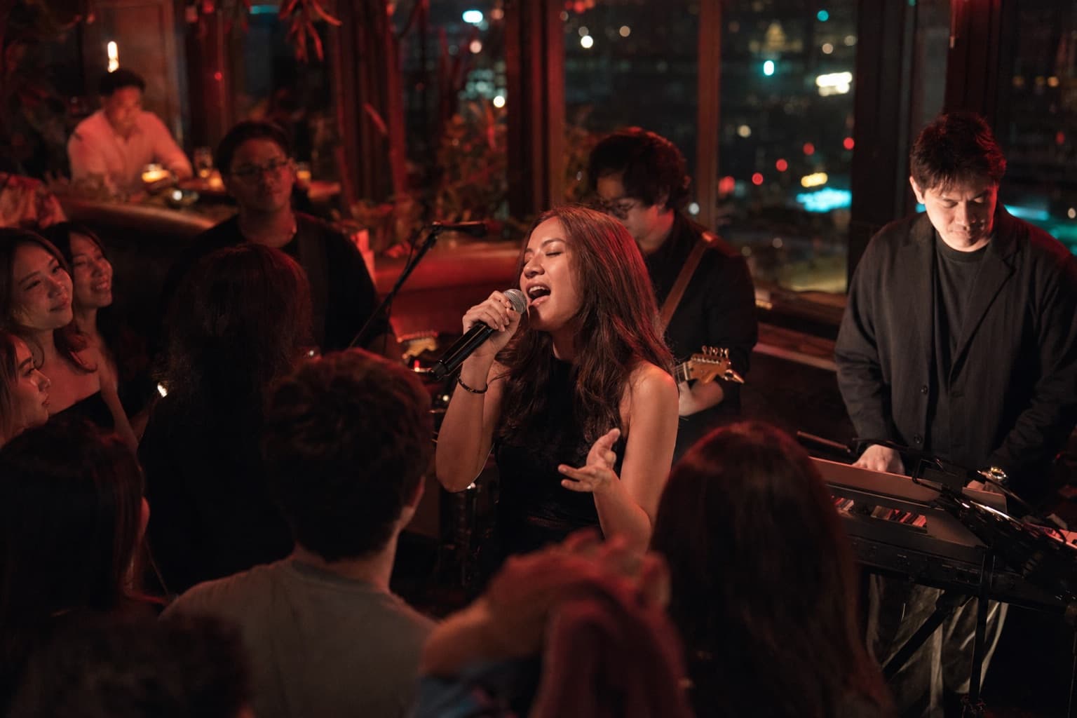 A vocalist mid-song with her band, framed by a rooftop bar in Bangkok and a crowd close in around her.